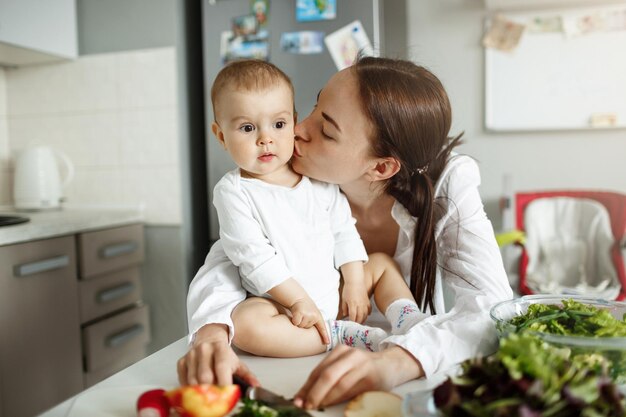 Portrait of happy beautiful mother kissing her lovely baby in cheek in dinner room Baby sitting on table with surprised expression