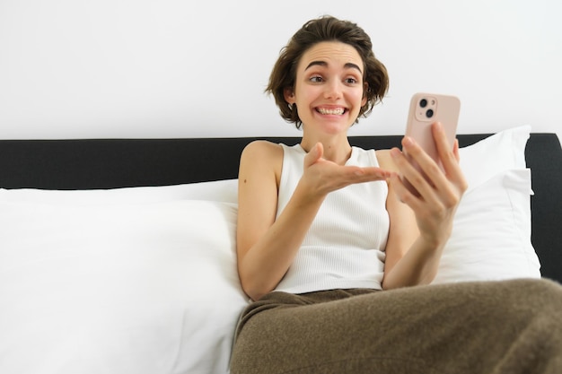 Free photo portrait of happy beautiful young woman in bed lying in bedroom and looking at smartphone chatting