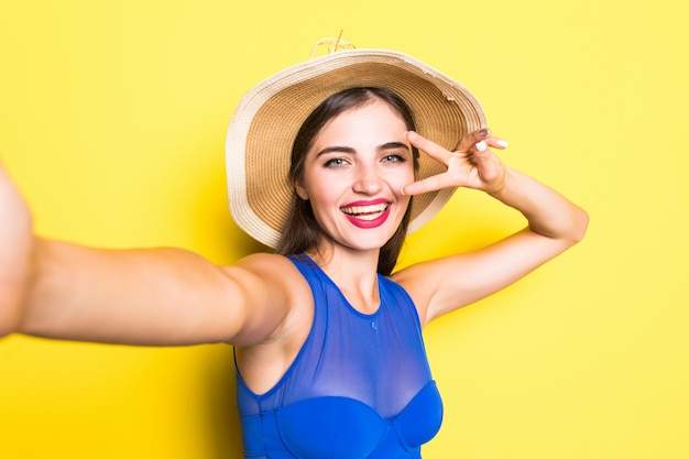 Ritratto di bella giovane donna felice che prende selfie sul suo telefono e che sorride, indossando costume da bagno in cappello di paglia sulla parete gialla