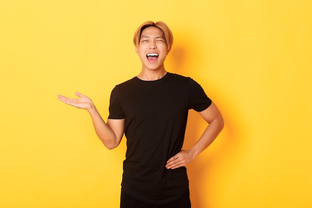 Portrait of happy blond asian guy, winking sassy and smiling, holding something on hand over yellow wall