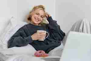 Free photo portrait of happy blond girl female model lying in bed in cosy clothes eating and watching movie on