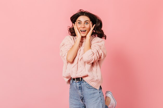 ピンクのセーターで幸せな青い目の女性の肖像画。巻き毛のブルネットの女性は、孤立した背景のカメラを喜んで覗きます。