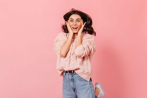 Portrait of happy blue-eyed lady in pink sweater. Curly brunette woman happily looks into camera on isolated background.