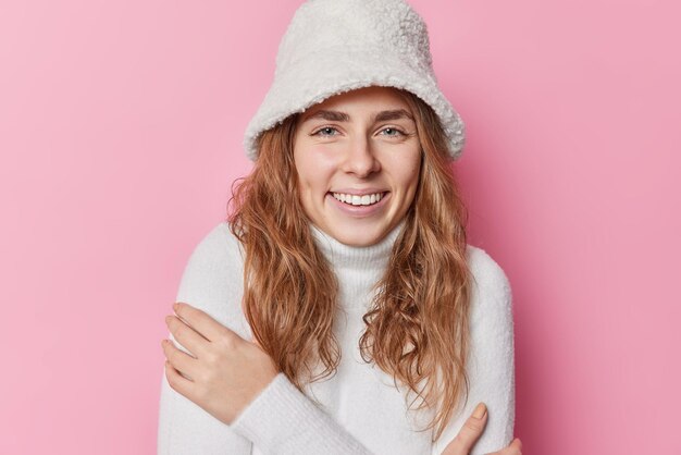 Portrait of happy blue eyed woman with long hair embraces herself smiles toothily wears white hat and jumper feels delighted isolated over pink background has natural beauty Emotions concept