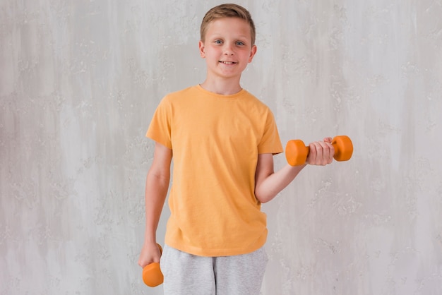 Ritratto di un ragazzo felice che si esercita con il dumbbell che si leva in piedi davanti al muro di cemento