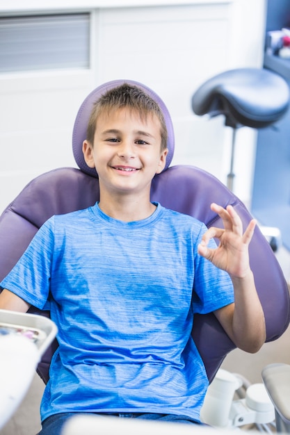 Foto gratuita ritratto di un ragazzo felice che si siede sulla sedia dentale che gesturing segno giusto