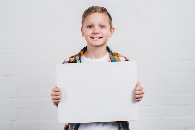 Портрет счастливого мальчика стоя против белой стены показывая белый пустой плакат
