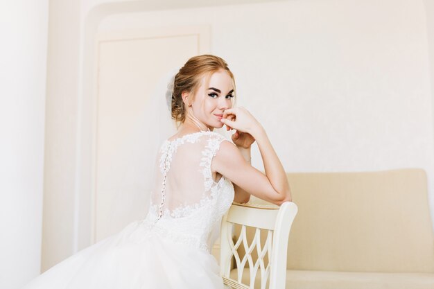 Portrait happy bride in wedding dress in appartment sitting on chair.