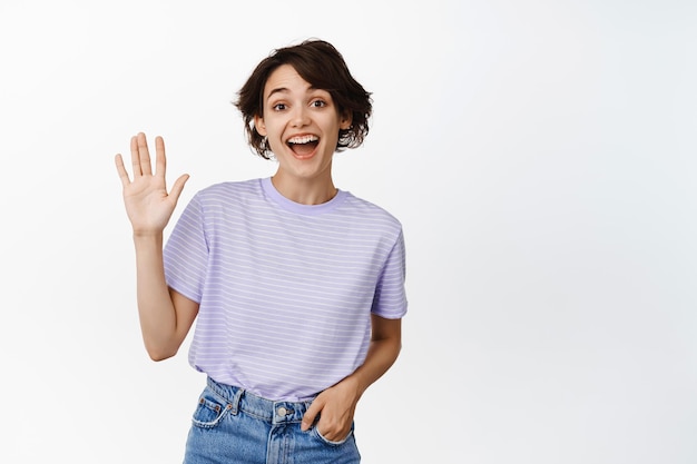 Portrait of happy brunette girl saying hello, waving at you and laughing, seeing a friend, greeting someone, standing in casual clothes against white background