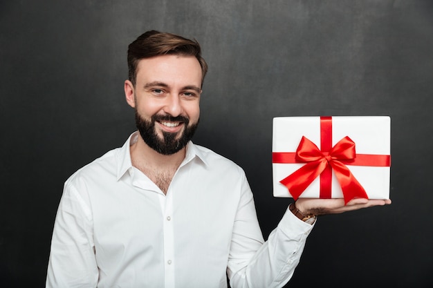 カメラに赤い弓と白いプレゼントボックスを示すと暗い灰色の壁に笑みを浮かべて幸せなブルネット男の肖像
