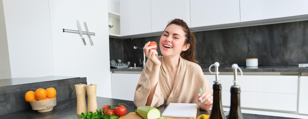 Free photo portrait of happy brunette woman bites tomato sits in the kitchen with vegetables cooking vegetarian