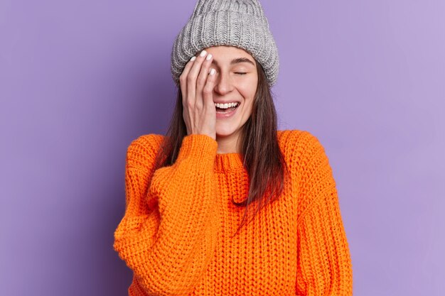 Портрет счастливой брюнетки закрывает лицо рукой, положительно хихикает, закрывает глаза, выражает положительные эмоции, носит вязаную шапку и свитер.