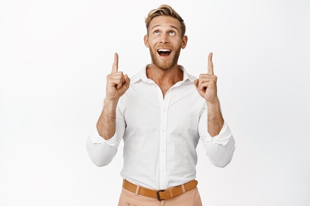 Portrait of happy businessman pointing and looking up with pleased smile checking out big discount standing over white background