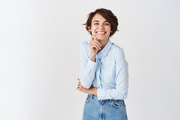 Portrait of happy candid woman smiling, looking cheerful and upbeat, touching face without makeup, standing on white wall