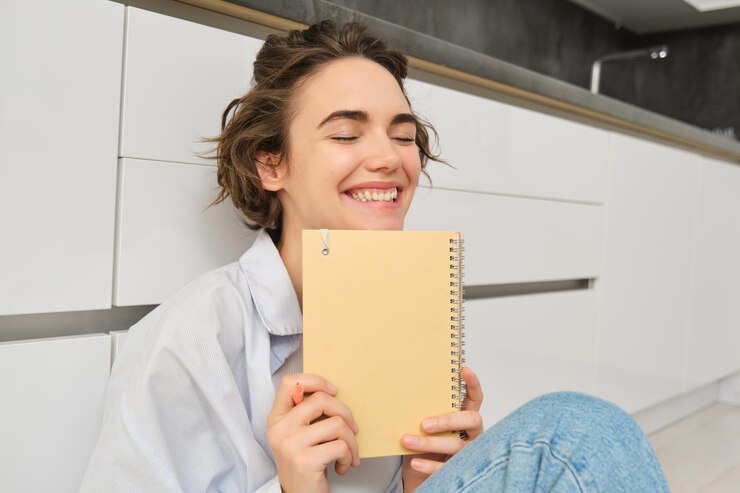 Free Photo portrait of happy carefree woman writes in her journal and laughs holds her diary reads something in