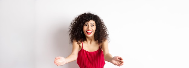 Free photo portrait of happy caucasian woman in red dress and makeup stretch out hands to beckon someone inviti