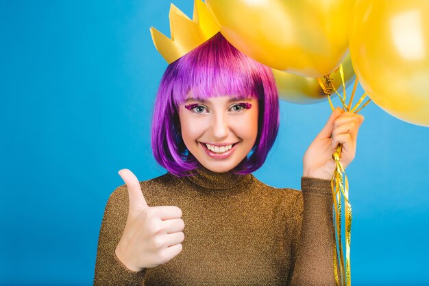 Portrait happy celebrating moments of joyful young woman with golden balloons smiling. Luxury dress, cut purple hair, princess crown, cheerful mood.