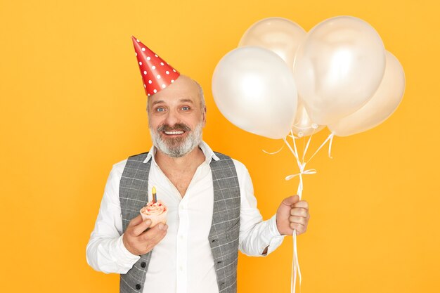 Ritratto di felice allegro anziano barbuto maschio che indossa abiti eleganti e cappello a cono in posa isolato holding compleanno cupcake ed elio palloncini