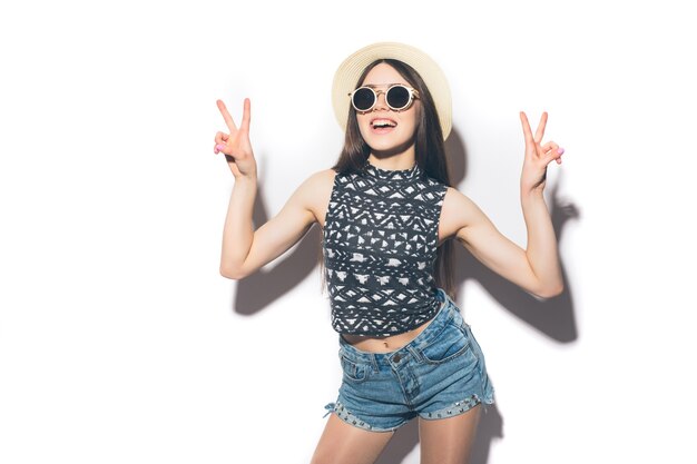 Portrait of a happy cheerful girl in summer hat showing peace gesture with two hands isolated