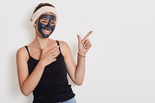 Portrait of happy cheerful woman improves her face skin, applies peeling mask, being in high spirit, models posing against white wall and pointing with both hands aside.