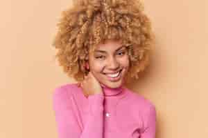 Free photo portrait of happy cheerful woman keeps hand on neck smiles gladfully has natural curly hair healthy skin feels happy wears pink poloneck isolated over brown background. people and emotions concept