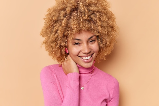 Portrait of happy cheerful woman keeps hand on neck smiles gladfully has natural curly hair healthy skin feels happy wears pink poloneck isolated over brown background. People and emotions concept