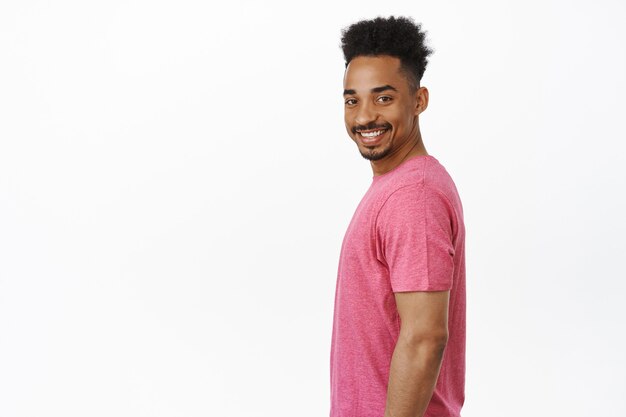 Портрет счастливого и уверенного в себе афро-американского парня, стоящего в профиль, повернувшего голову с большой веселой улыбкой на лице, стоя в розовой футболке на белом