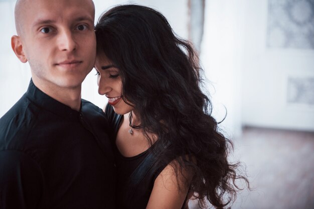 Portrait of happy couple indoors. Bald guy and brunette woman stands in the white room