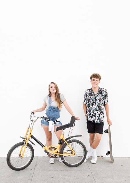 Portrait of a happy couple standing with bicycle and skateboard in front of wall