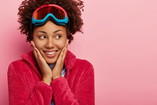 Portrait of happy curly haired girl snowboader enjoys active rest, touches cheeks gently, looks away with toothy smile, wears red coat, ski glasses, isolated on pink background.