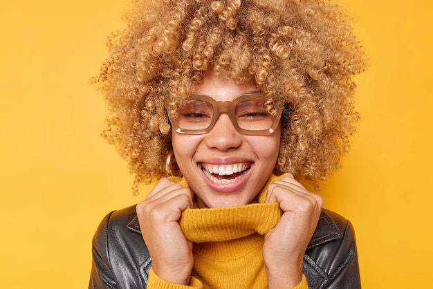 Portrait of happy curly haired woman keeps hands on collar of jumper wears leather jacket and sweater smiles broadly shows white teeth isolated over yellow background Positive emotions concept