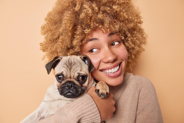 幸せな縮れ毛の女性の肖像画ペットの飼い主は、ベージュの背景の上に分離されたカジュアルなジャンパーを喜んで身に着けているお気に入りの子犬の笑顔を抱きしめるパグ犬への愛とケアを表現しています。感情的な態度