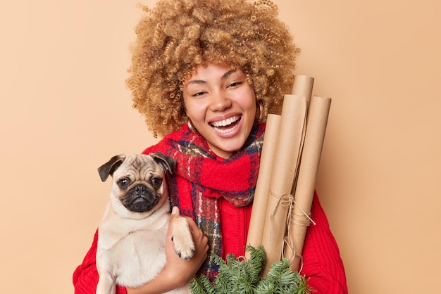 血統の犬と新年の装飾の笑顔で幸せな縮れ毛の女性ペットの所有者のポーズの肖像画は、茶色の背景の上に分離された冬の服に身を包んだポジティブなお祭り気分を持っています