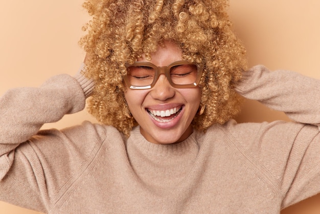 Free photo portrait of happy curly haired young woman keeps hands behind head laughs joyfully keep eyes closed wears spectacles and casual jumper isolated over brown background positive emotions concept