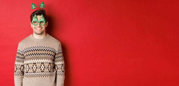 Portrait of happy and cute man in winter sweater and party glasses celebrating new year or christmas smiling while standing over red background