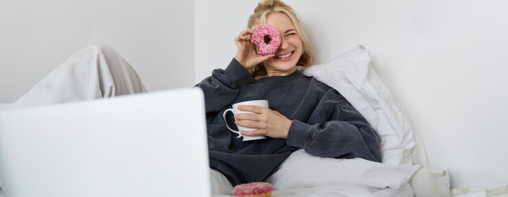 Free Photo portrait of happy cute woman lying in bed with laptop drinking tea looking through doughnut hole and