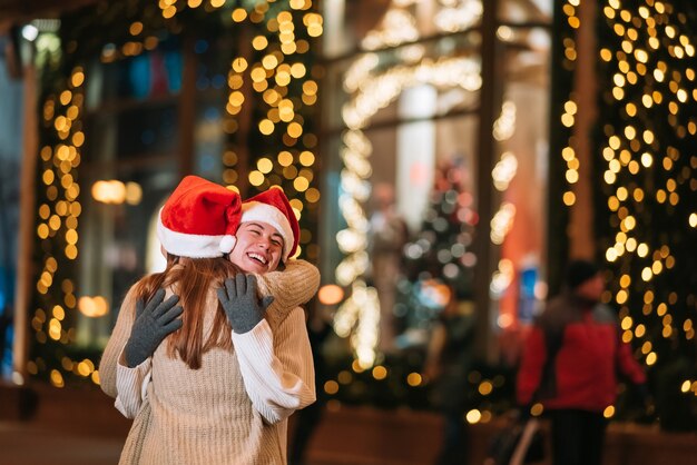 Портрет счастливых милых молодых друзей, обнимающих друг друга и улыбающихся во время прогулки в канун Рождества на открытом воздухе.