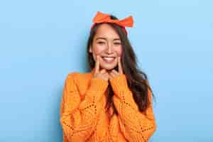 Free photo portrait of happy dark haired korean girl with toothy smile, keeps index fingers near mouth, wears orange headband and knitted sweater