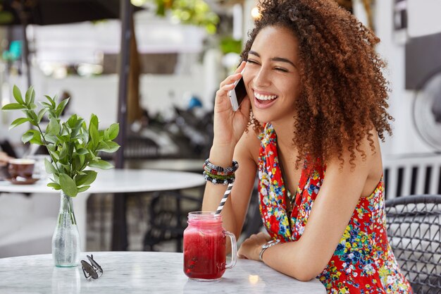 スマートフォンで友だちとコミュニケーションをとりながら、真っ黒に笑う幸せな黒肌の女性の肖像画は、コーヒーショップで自由な時間を過ごしています。