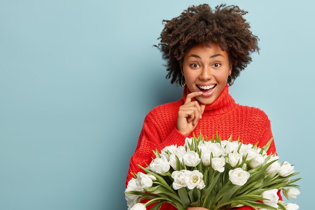 Portrait of happy dark skinned girl with curly hair, smiles joyfully, keeps finger on lower lip, dressed in red sweater, enjoys spring time, holds white tulips, isolated on blue wall