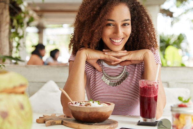 Free photo portrait of happy dark skinned young female with frizzy hair, eats something and drinks smoothie, spends free time with boyfriend or friend, enjoys summer vacations in tropical country on island