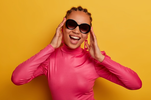Portrait of happy dark skinned young woman wears trendy sunglasses and pink sweater, enjoys sunny day, dressed in bright clothes, isolated on yellow wall. People, fashion and style concept.