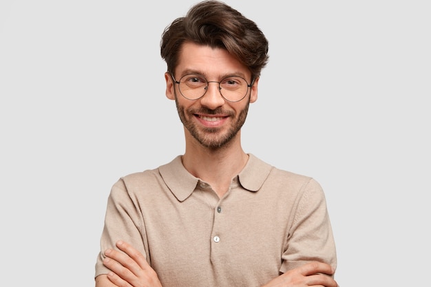 Portrait of happy delighted attractive man keeps hands crossed, dressed in casual outfit, looks joyfully, being self confident in success, isolated over white wall