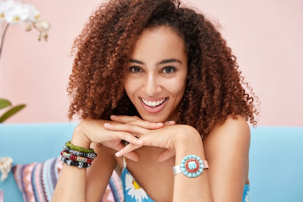 Free photo portrait of happy delighted dark skinned mixed race female with frizzy bushy hair, feels relaxed as sits on comfortable sofa against pink wall