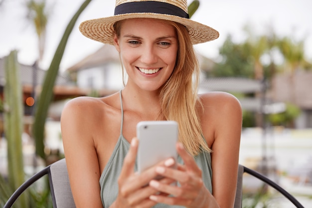 Портрет счастливой, довольной женщины в соломенной шляпе, с веселым выражением лица, читает что-то на смартфоне, проводит свободное время в дорогом летнем кафе, отдыхает летом. Улыбающаяся женщина с мобильным телефоном