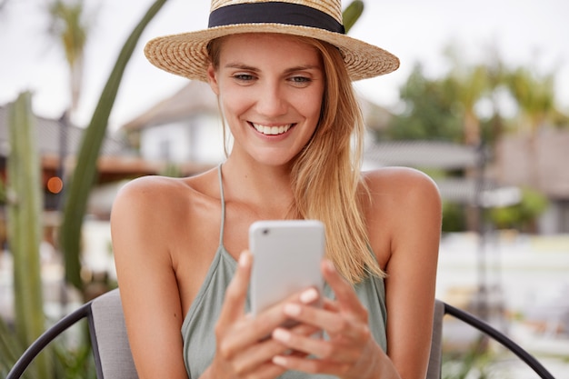 Portrait of happy delighted female wears straw hat, has cheerful expression, reads something on smart phone, enjoys free time in expensive outdoor cafe, has summer rest. Smiling woman with mobile