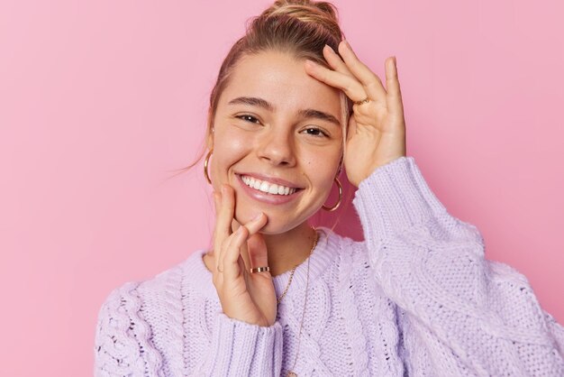 Portrait of happy delighted woman touches face getly has healthy clean skin smiles toothily wears casual knitted sweater isolated over pink background Charming female model expresses sincere feelings