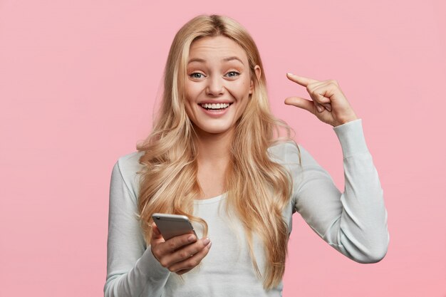 Portrait of happy delighted young cute female shows ammount of money she recieved this month, keeps modern cell phone in hands, reads message, isolated over pink wall. This is too small