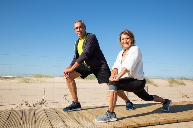 Портрет счастливой пожилой пары, вместе занимающейся тренировкой. Кавказский мужчина и женщина в спортивной одежде полусидя, растягивая ноги, улыбаясь, глядя в камеру. Свободное время для занятий спортом на пенсии