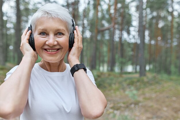 portrait of happy energetic middle aged woman listening to music while jogging outdoors, holding hands on headphones